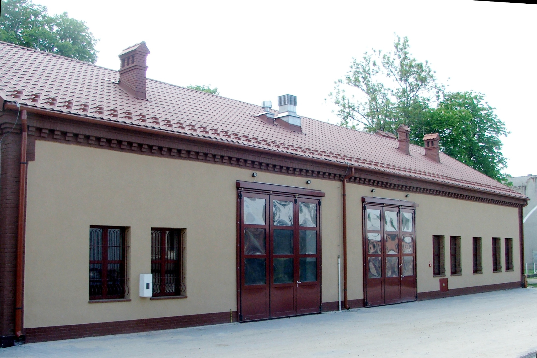 Parterowy budynek z beżową elewacją, czerwoną dachówką i dużymi, przeszklonymi, dwuskrzydłowymi drzwiami garażowymi.