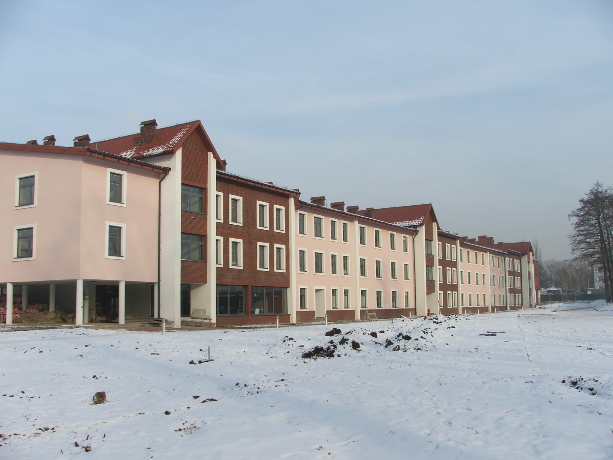 Wielorodzinny budynek mieszkalny w trakcie budowy, z widocznymi elementami konstrukcyjnymi, częściowo w kolorze cegły, częściowo otynkowany na różowo, z czerwonym dachem, pokryty śniegiem,...