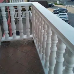 Biała balustrada balkonowa z toczonymi tralkami, widok z góry na brązową terakotę i zaparkowane samochody w tle.