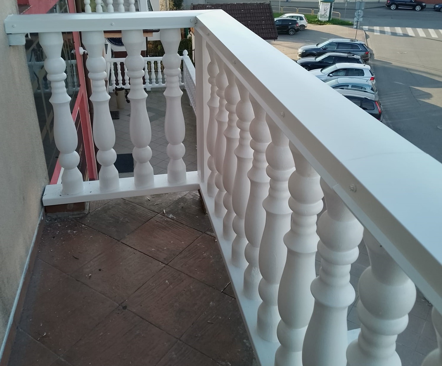 Biała balustrada balkonowa z toczonymi tralkami, widok z góry na brązową terakotę i zaparkowane samochody w tle.