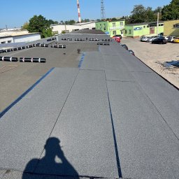 Sebastian Lewanowski - Szary, płaski dach pokryty papą, z widocznymi rolkami papy w tle i cieniem osoby robiącej zdjęcie. W tle budynki i komin.