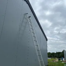 Szara ściana budynku obita blachą trapezową z drabiną opartą o ścianę, widok z dołu, pochmurne niebo w tle.