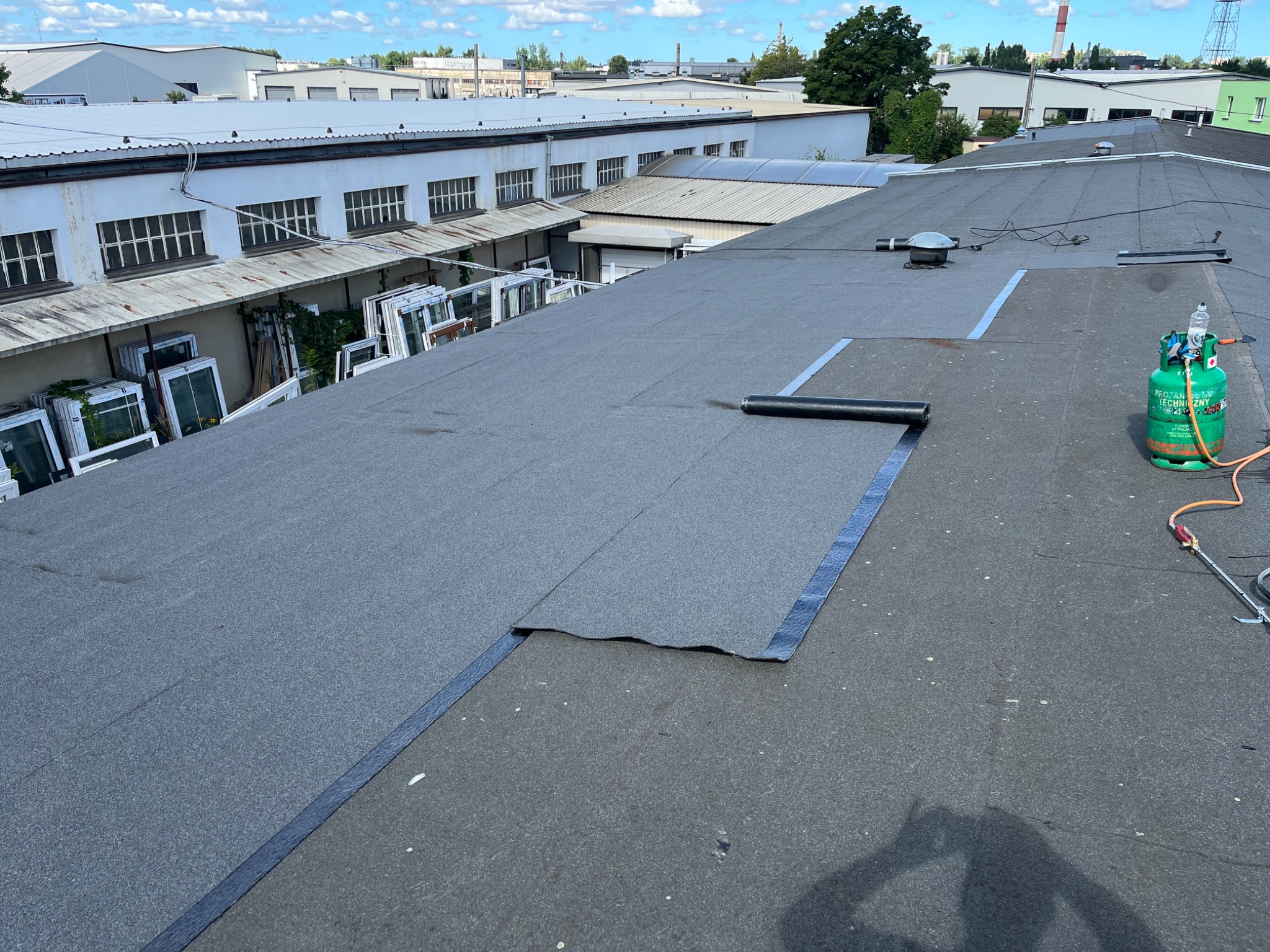 Bitumiczna papa termozgrzewalna na dachu budynku przemysłowego, częściowo ułożona. Widoczna butla z gazem i rolka papy. W tle budynki i błękitne niebo.