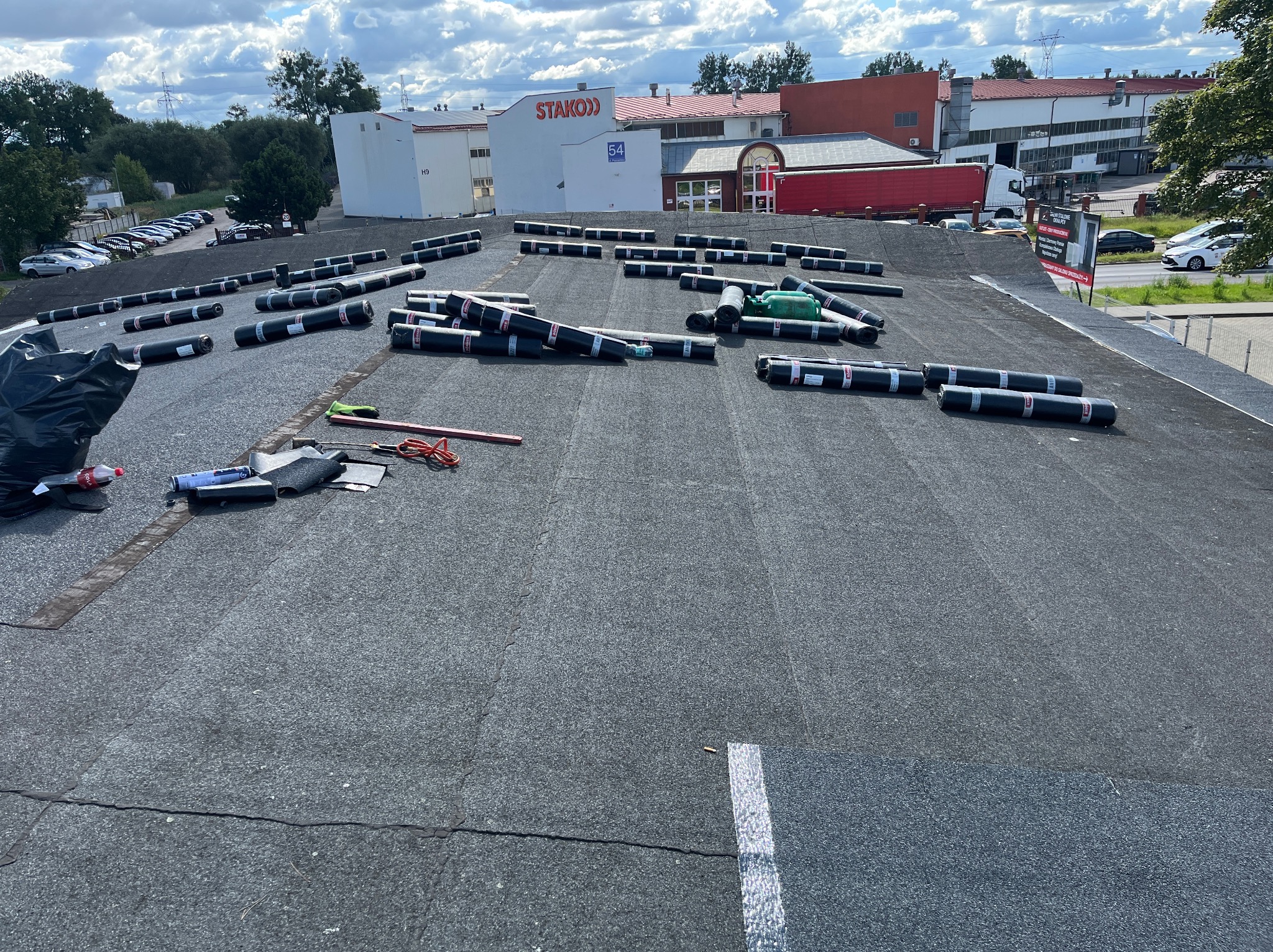 Przygotowanie do krycia dachu papą termozgrzewalną. Widoczne rolki papy, narzędzia dekarskie i fragment dachu z widokiem na budynek Stako i zaparkowane samochody.