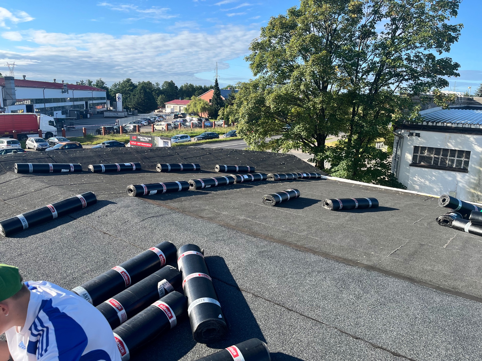 Dach pokryty papą termozgrzewalną w rolkach marki Börner. Widoczny fragment sylwetki dekarza w trakcie pracy. W tle zabudowania miejskie i błękitne niebo.