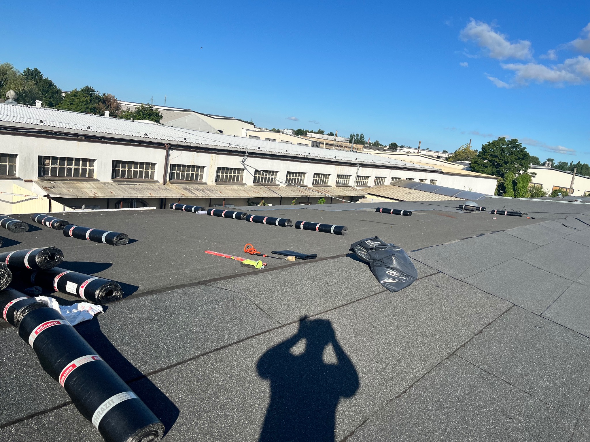 Prace dekarskie w Słupsku: czarny, płaski dach pokryty papą termozgrzewalną. Widoczne rolki papy i narzędzia dekarskie. Budynki w tle i błękitne niebo.