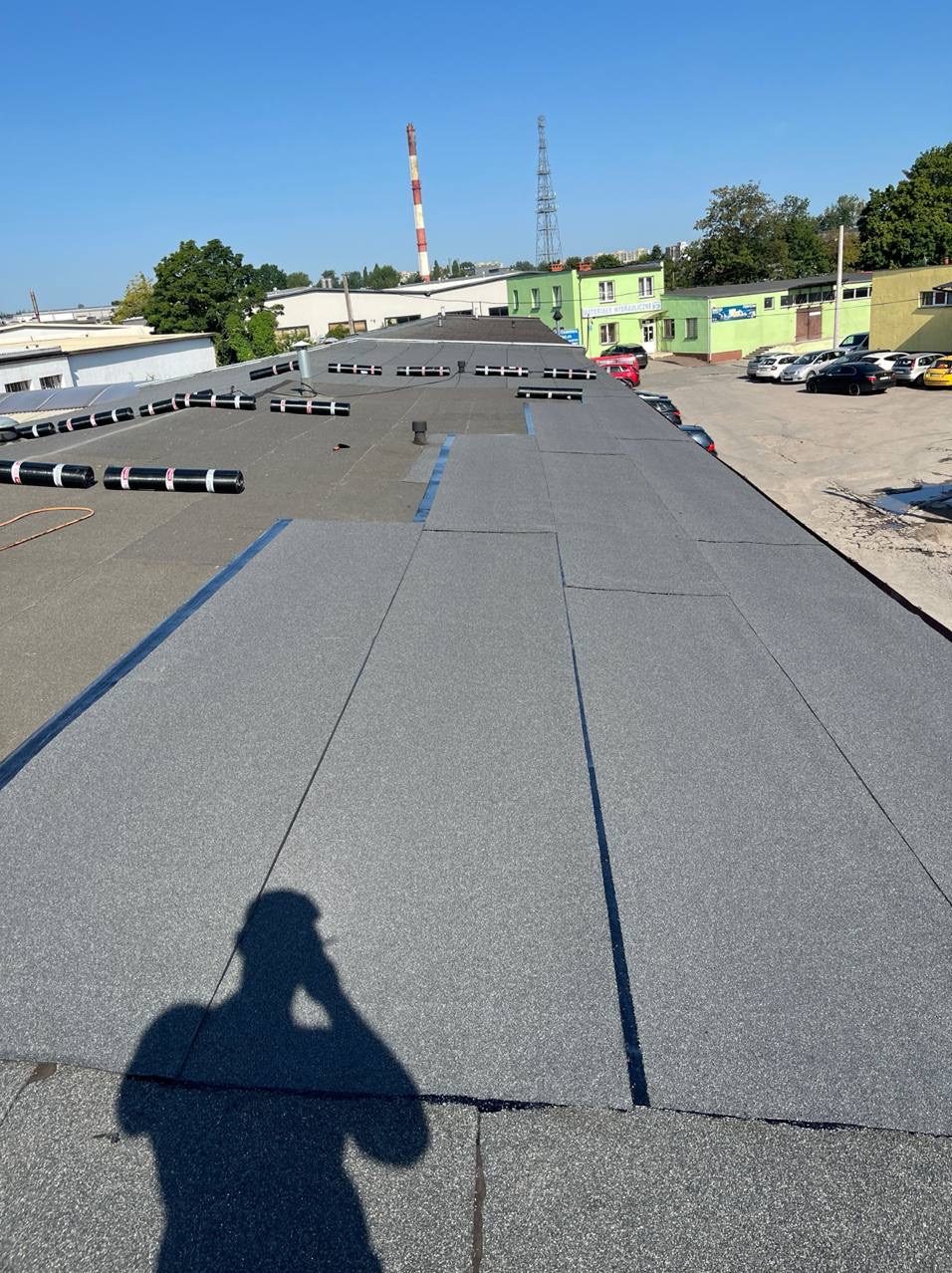 Szary, płaski dach pokryty papą termozgrzewalną z widocznym cieniem osoby robiącej zdjęcie. W tle budynki przemysłowe i komin.