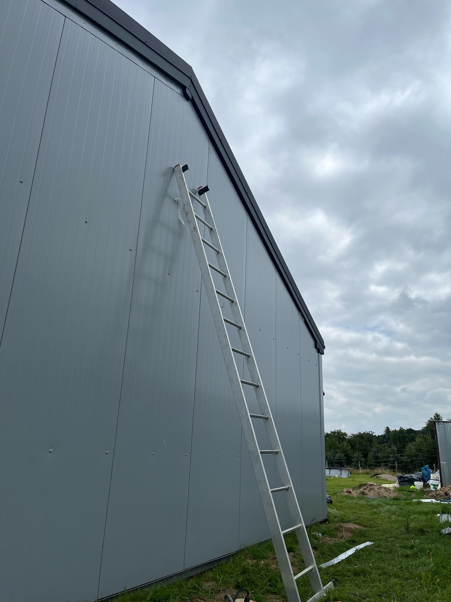 Szara ściana budynku obita blachą trapezową z drabiną opartą o ścianę, widok z dołu, pochmurne niebo w tle.