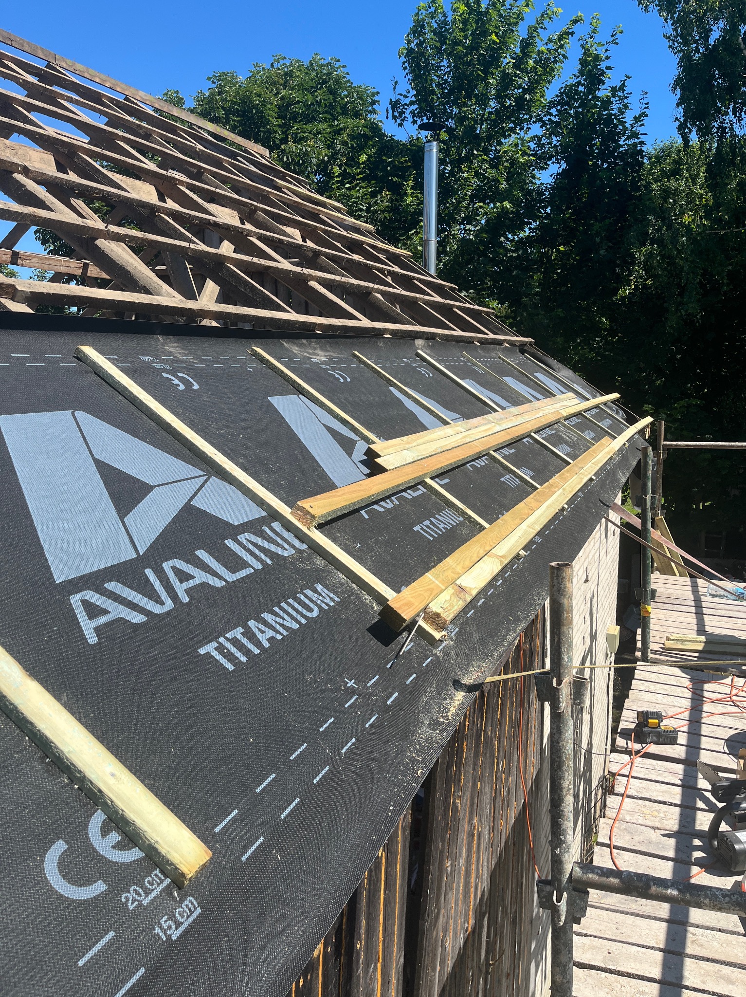 Dach z widoczną konstrukcją drewnianą i membraną Avaline Titanium, zabezpieczoną listwami. W tle zieleń drzew i błękit nieba. Widoczne rusztowanie i narzędzia.