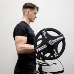 Młody mężczyzna w czarnej koszulce, z bicepsem napiętym podczas ćwiczenia z czarnym obciążeniem 15 kg marki PROUD, na tle białej ściany z czarnym rysunkiem muskularnej sylwetki.