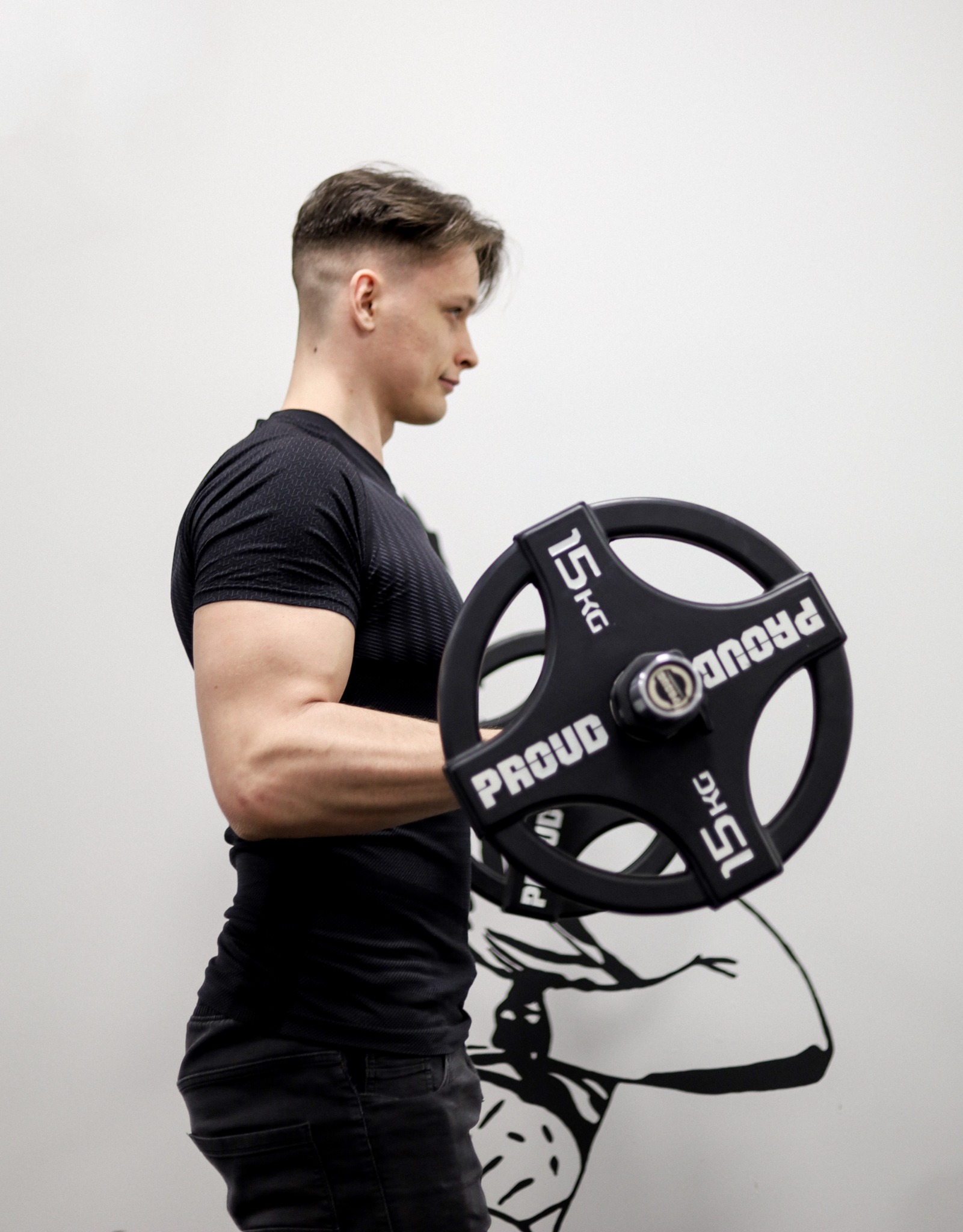 Młody mężczyzna w czarnej koszulce, z bicepsem napiętym podczas ćwiczenia z czarnym obciążeniem 15 kg marki PROUD, na tle białej ściany z czarnym rysunkiem muskularnej sylwetki.