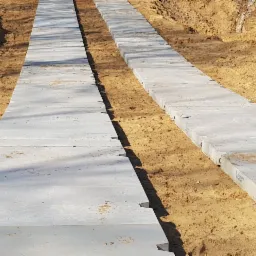 Dwa rzędy ułożonych, prefabrykowanych płyt betonowych w wykopie z piaszczystą ziemią, widoczne elementy łączące płyty, jasne światło słoneczne.