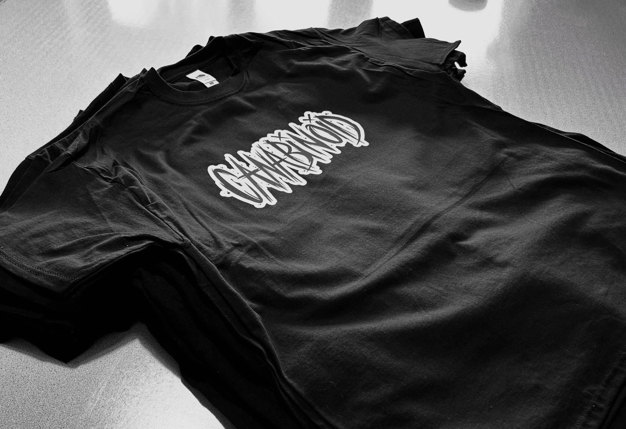 Stos czarnych T-shirtów z białym, unikalnym nadrukiem graficznym, ułożonych na jasnej powierzchni. Monochromatyczna tonacja podkreśla detale wzoru.