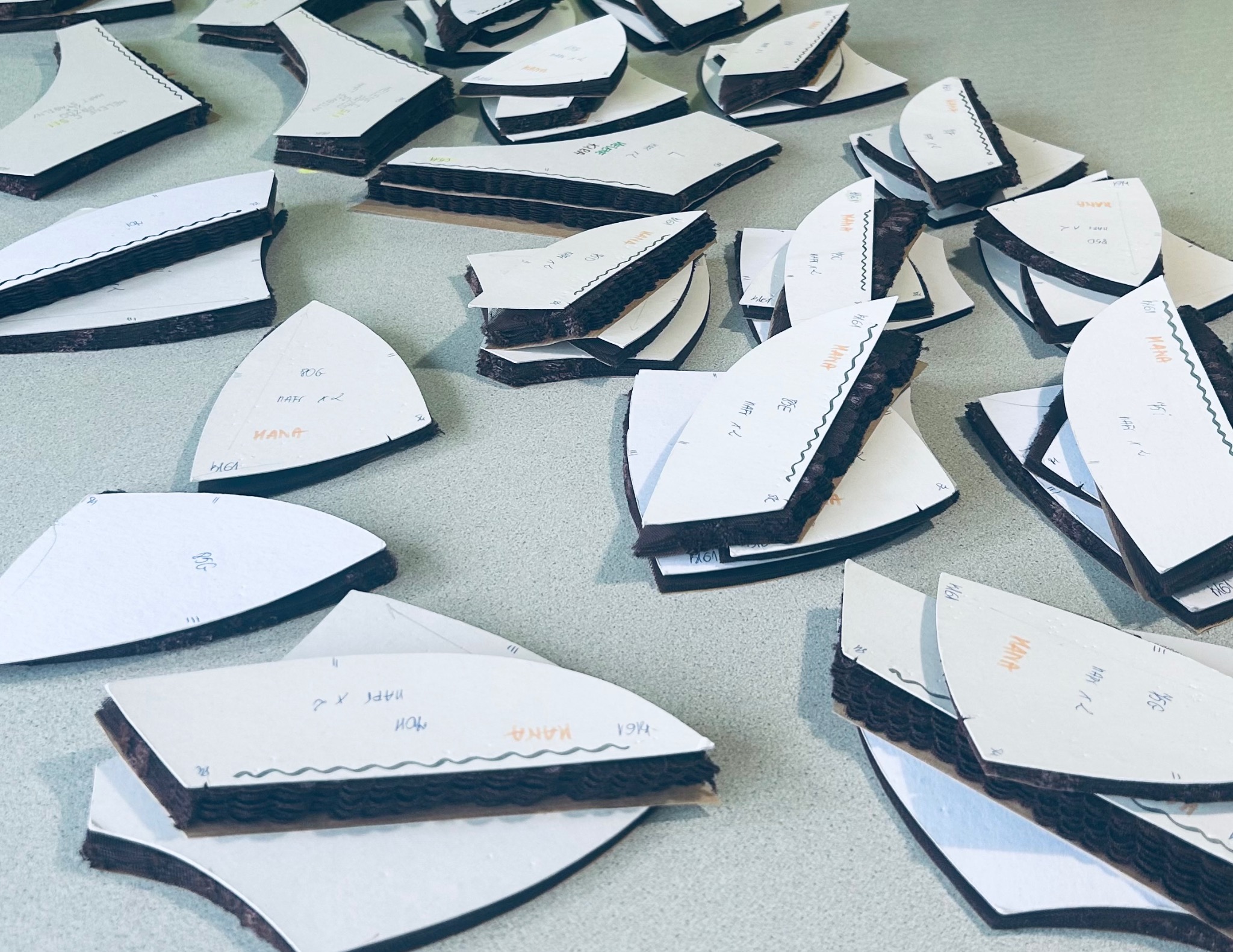 Wykroje odzieży ułożone na stole, przygotowane do cięcia. Widoczne warstwy materiału i papierowe szablony z naniesionymi oznaczeniami. Różne kształty i rozmiary wykrojów.