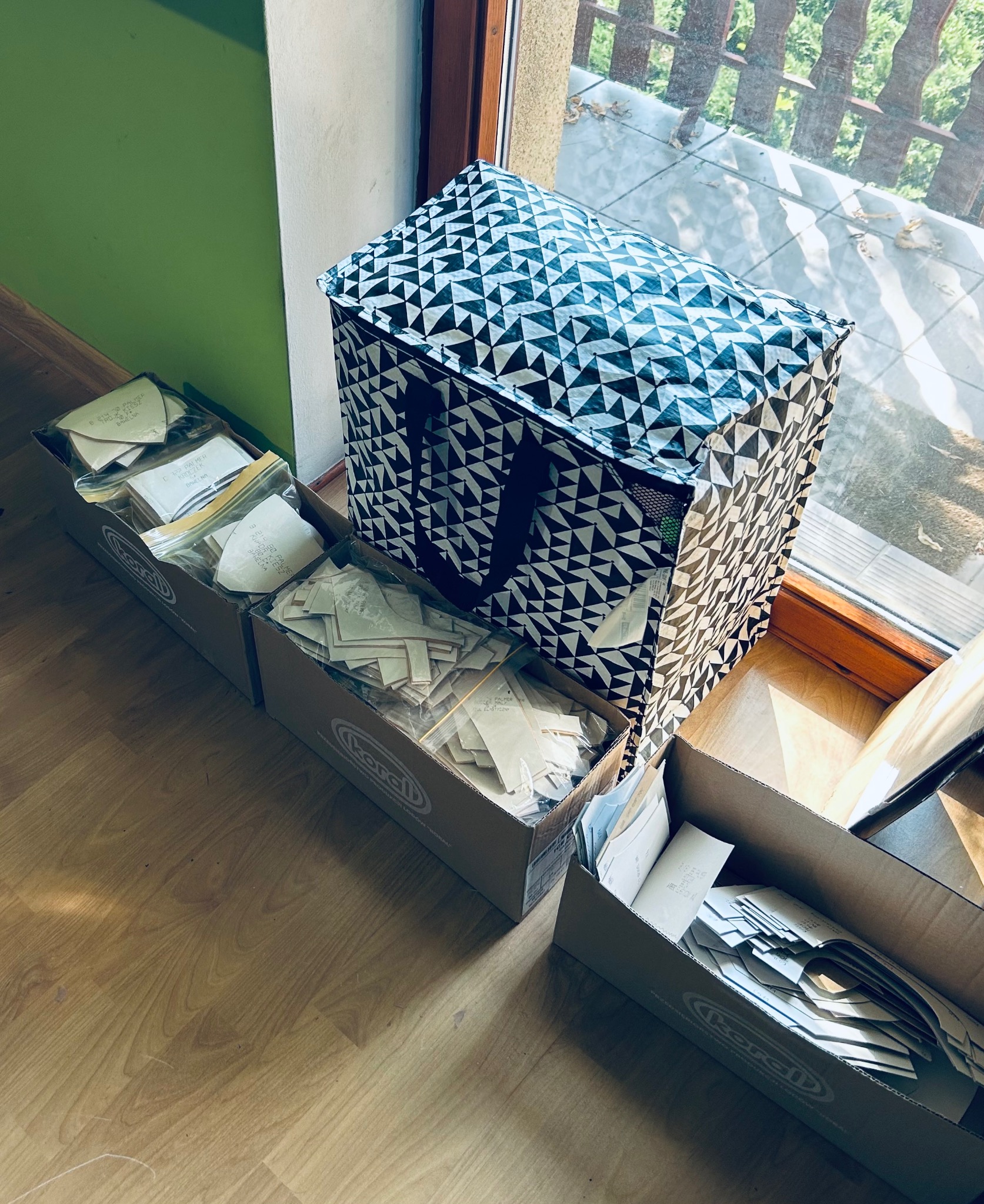Kroje odzieży w kartonach i torba z geometrycznym wzorem, ustawione na podłodze w pobliżu okna. Widoczne etykiety i wzory na papierze.