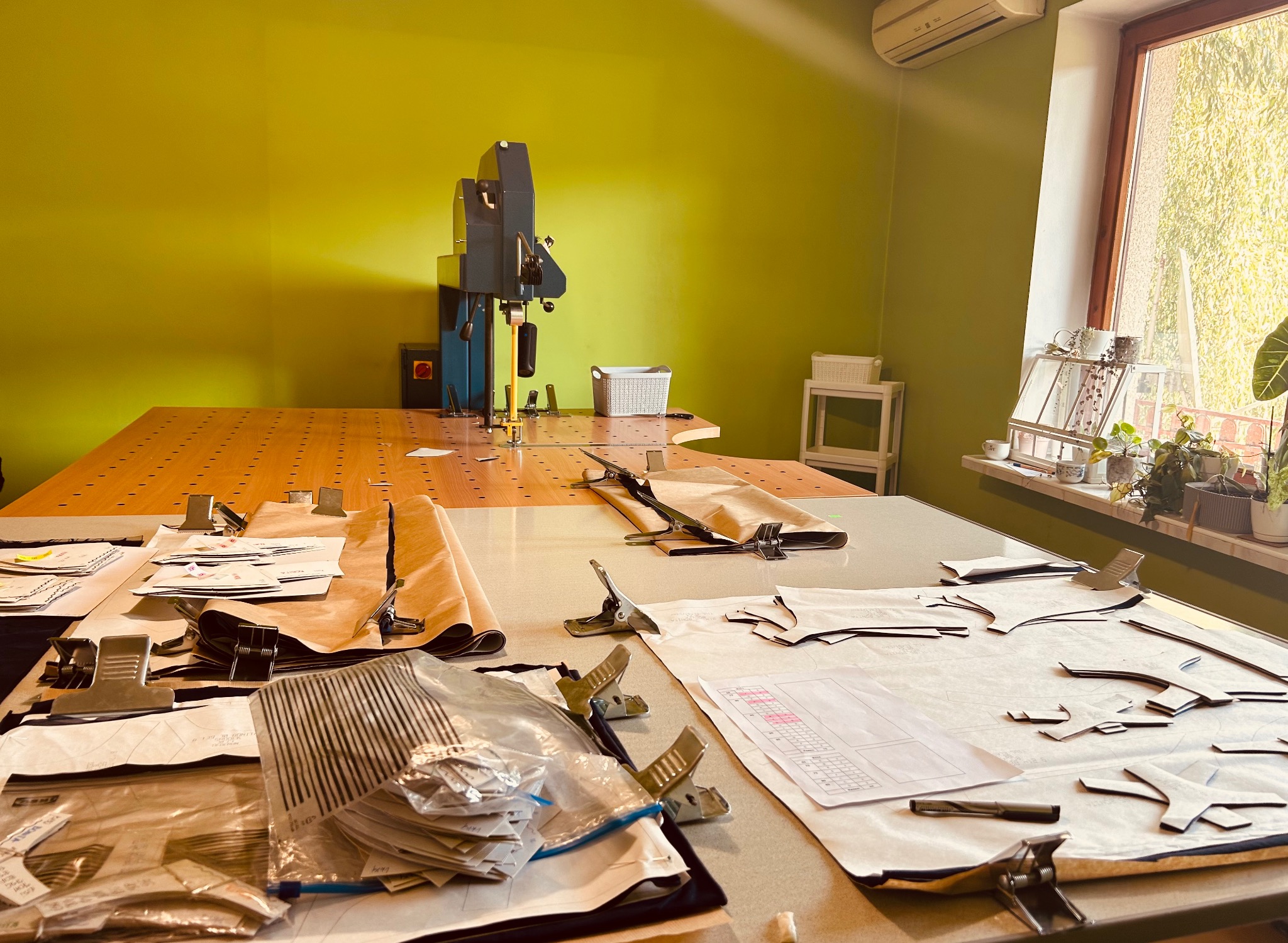 Duży stół krojczy z wykrojami, materiałami i maszyną do cięcia w tle. Widoczne detale: metalowe klipsy, wzory na papierze, elementy odzieży. Jasne, naturalne światło.