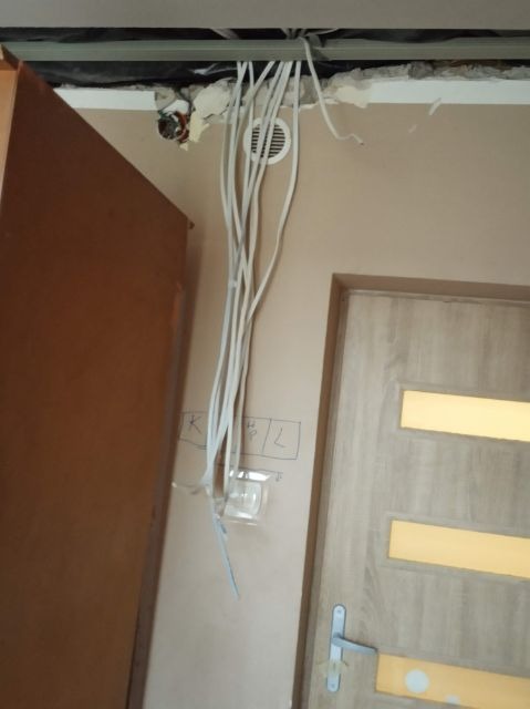 Wiązka przewodów elektrycznych wychodząca z sufitu w trakcie montażu, widoczne oznaczenia na ścianie, obok futryna drzwi wewnętrznych i fragment szafy.