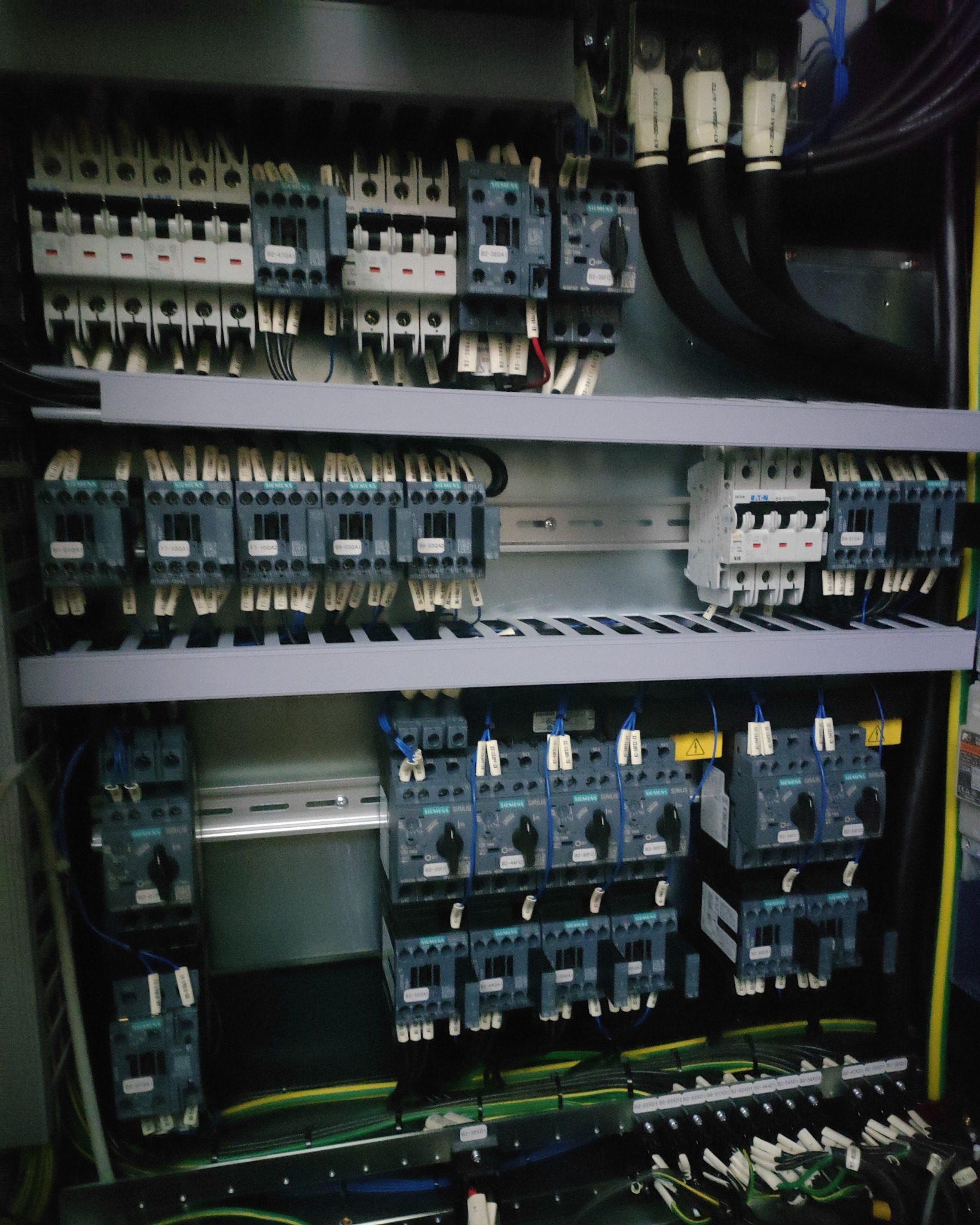 Rozdzielnica elektryczna z zamontowanymi bezpiecznikami, stycznikami i przewodami oznaczonymi etykietami, widoczne elementy marki Siemens i Eaton.