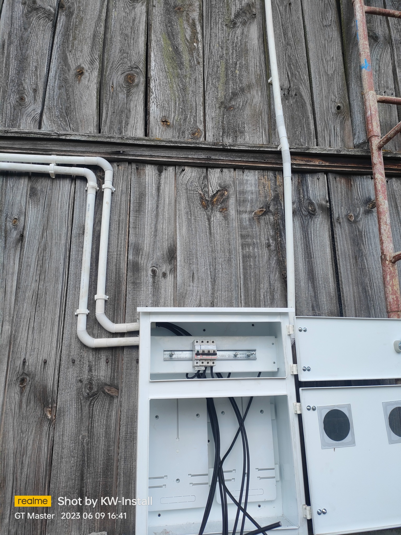 Otwarta skrzynka elektryczna z podłączonymi przewodami na tle drewnianej ściany i rur instalacyjnych