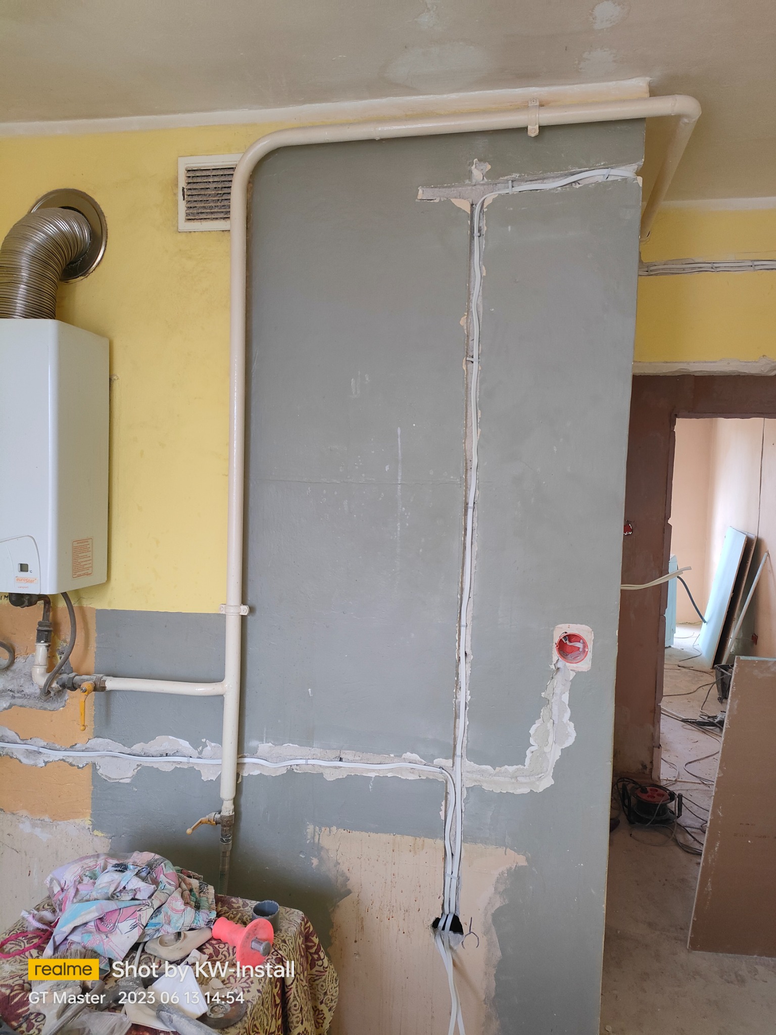 Ściana w trakcie remontu z widocznymi przewodami elektrycznymi w bruzdach, rurami instalacji grzewczej i wiszącym piecem gazowym, przygotowana do dalszych prac wykończeniowych.