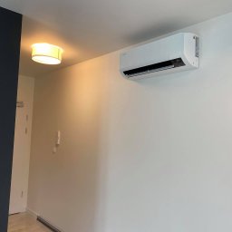 Climatis - Biała jednostka klimatyzacji Samsung zamontowana na jasnej ścianie w nowoczesnym wnętrzu, widoczna lampa sufitowa i fragment ściany w kolorze granatowym.