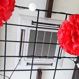 Climatis - Biała, sufitowa jednostka klimatyzacyjna z widocznymi kratkami wentylacyjnymi, udekorowana czerwonymi i białymi pomponami oraz lampkami, zamontowana pod drewnianą belką.