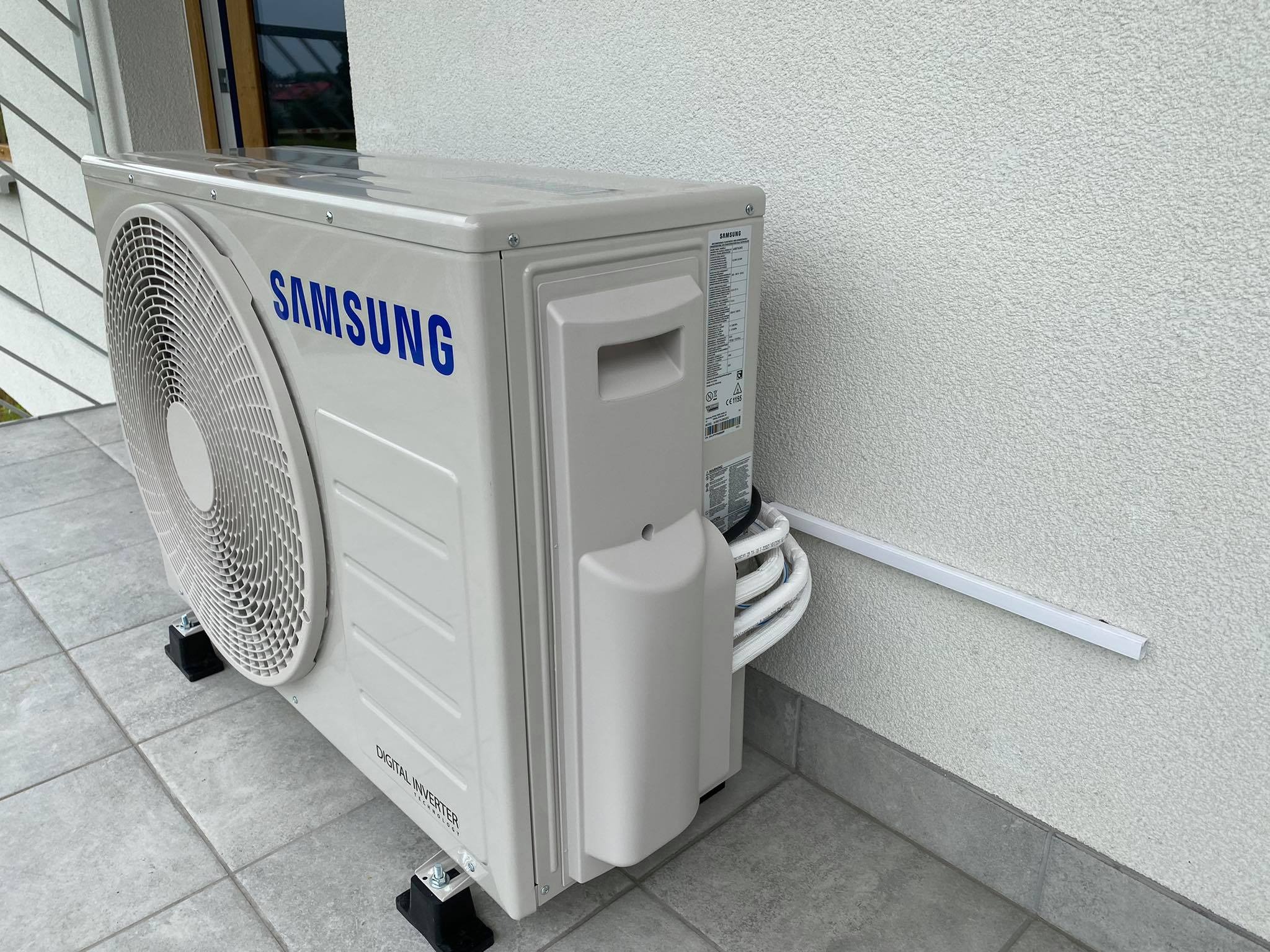 Zewnętrzna jednostka klimatyzacji Samsung Digital Inverter zamontowana na szarych płytkach, z widocznymi przewodami zabezpieczonymi listwą przyścienną.