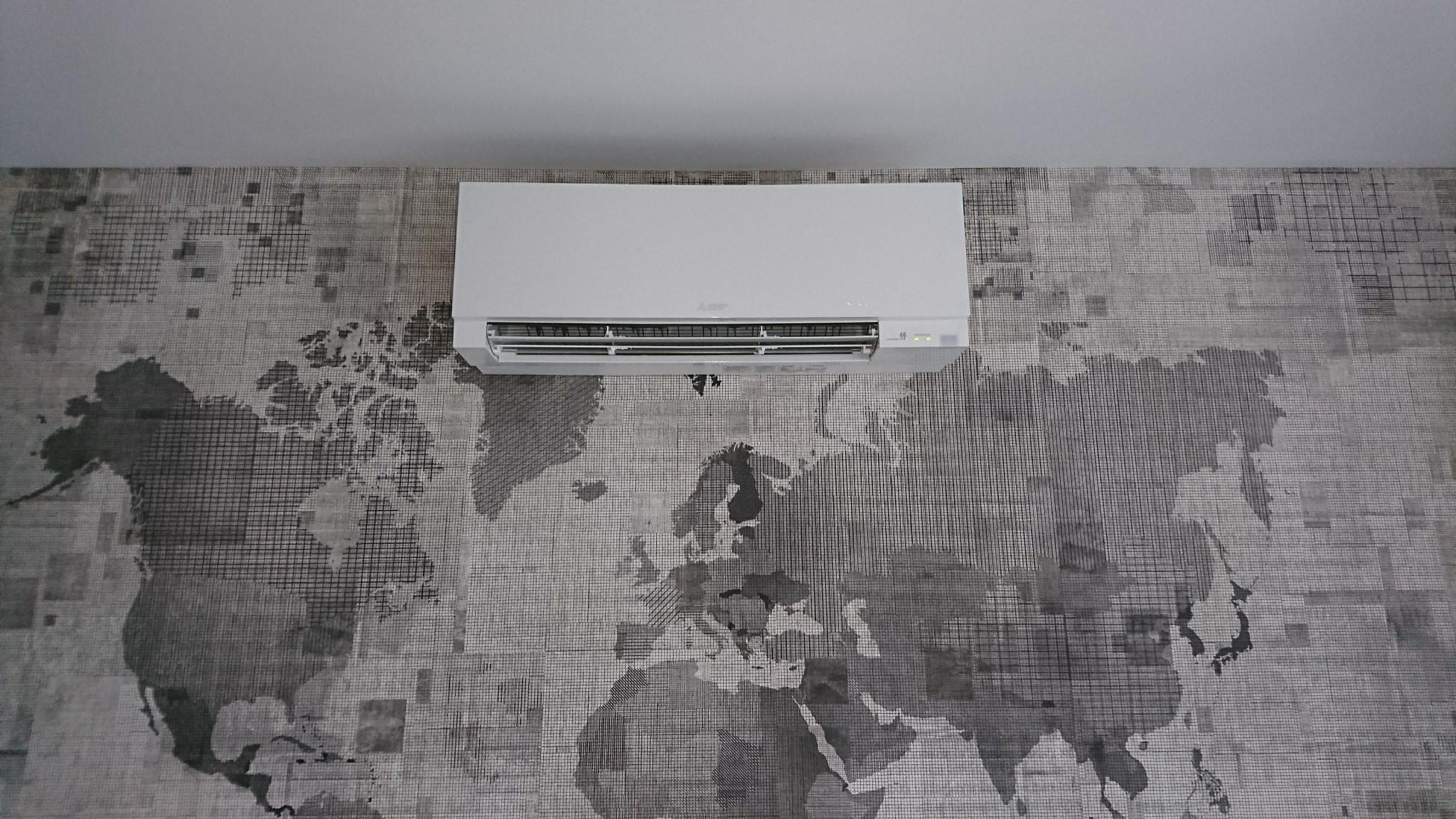 Biały klimatyzator ścienny zamontowany na ścianie z tapetą przedstawiającą mapę świata w odcieniach szarości.