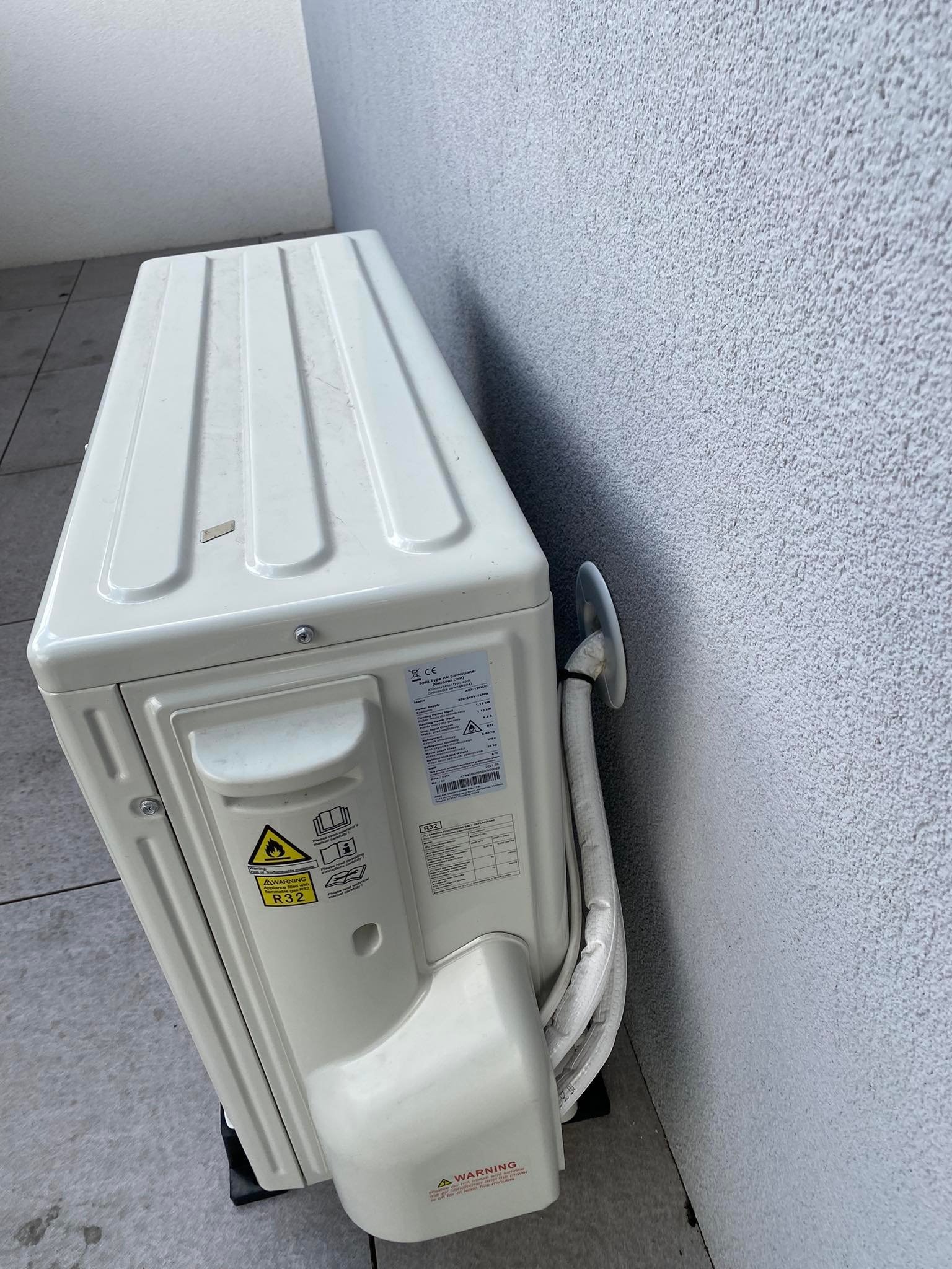 Zewnętrzna jednostka klimatyzacji typu split, zamontowana na szarej elewacji, widoczne przewody chłodnicze w białej osłonie oraz tabliczka znamionowa z informacjami technicznymi.
