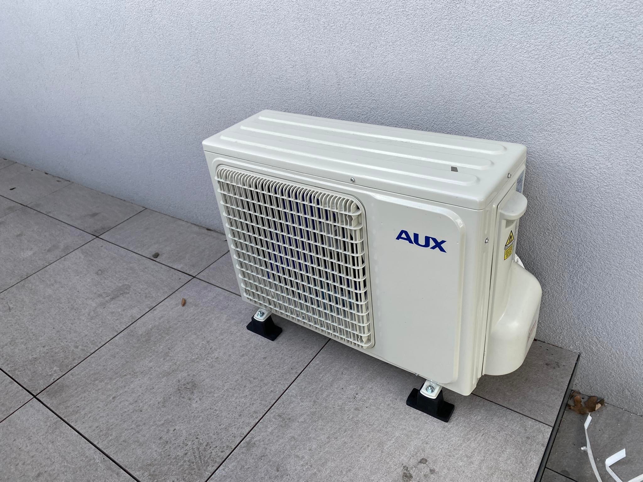 Zewnętrzna jednostka klimatyzacji marki AUX, umieszczona na szarym, płytkowym podłożu przy jasnej, teksturowanej ścianie budynku.