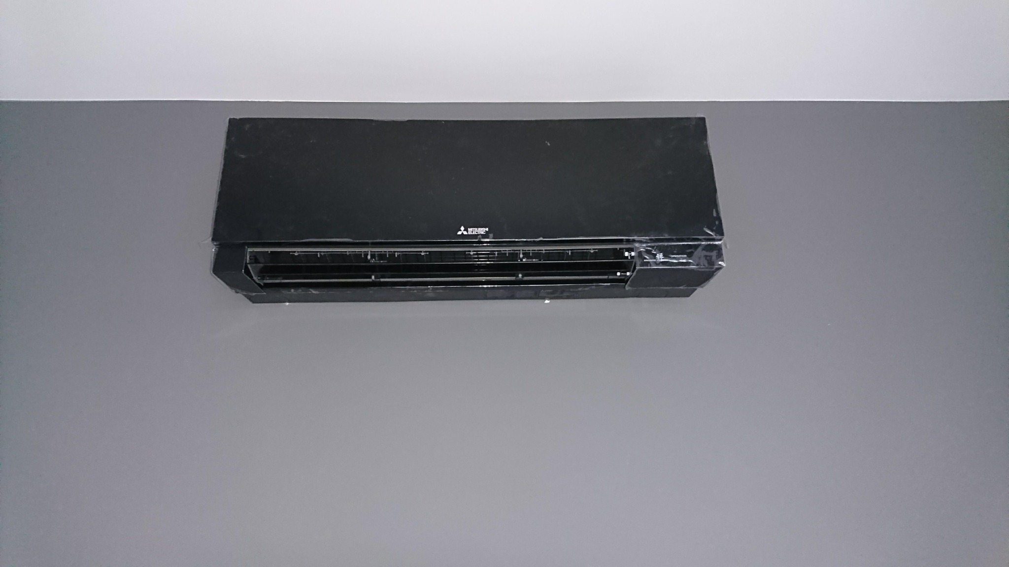 Czarna jednostka wewnętrzna klimatyzacji Mitsubishi, zamontowana na szarej ścianie pod białym sufitem, częściowo jeszcze w folii ochronnej.