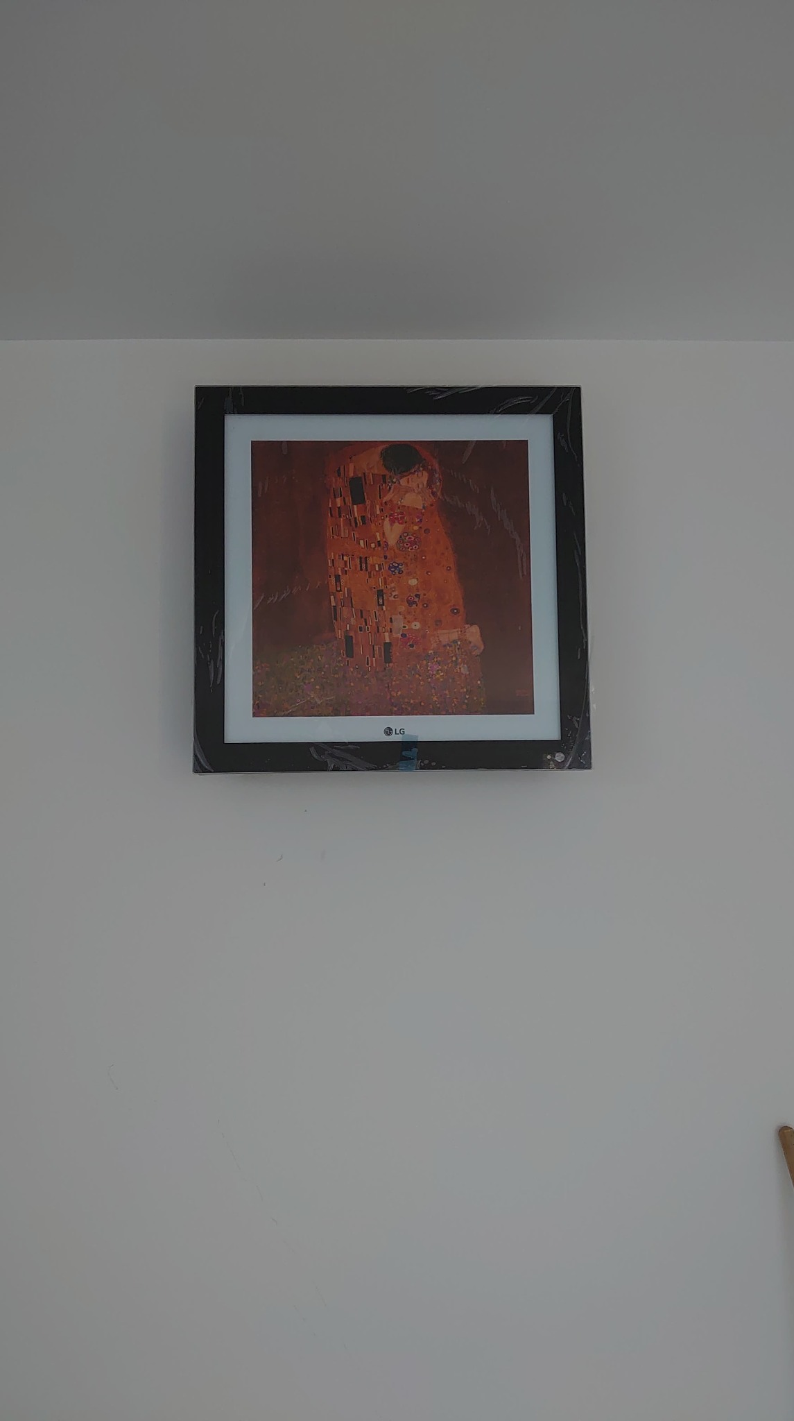Telewizor LG The Frame z wyświetloną reprodukcją obrazu 'Pocałunek' Gustava Klimta, zamontowany na białej ścianie, widoczne pozostałości folii ochronnej na ramie urządzenia.