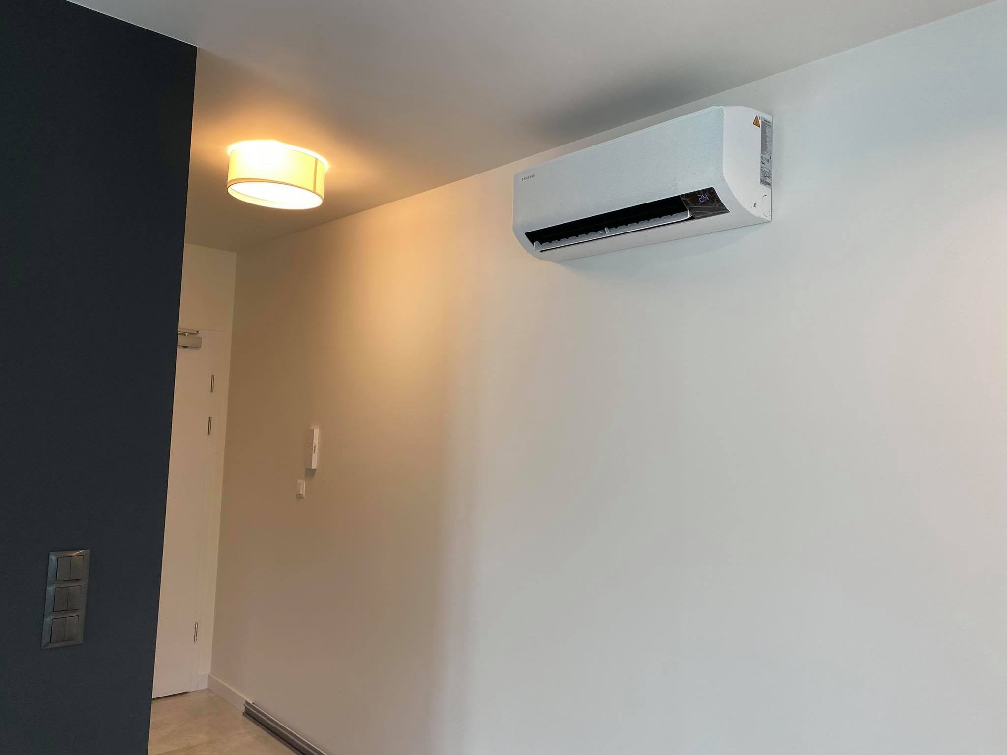 Biała jednostka klimatyzacji Samsung zamontowana na jasnej ścianie w nowoczesnym wnętrzu, widoczna lampa sufitowa i fragment ściany w kolorze granatowym.