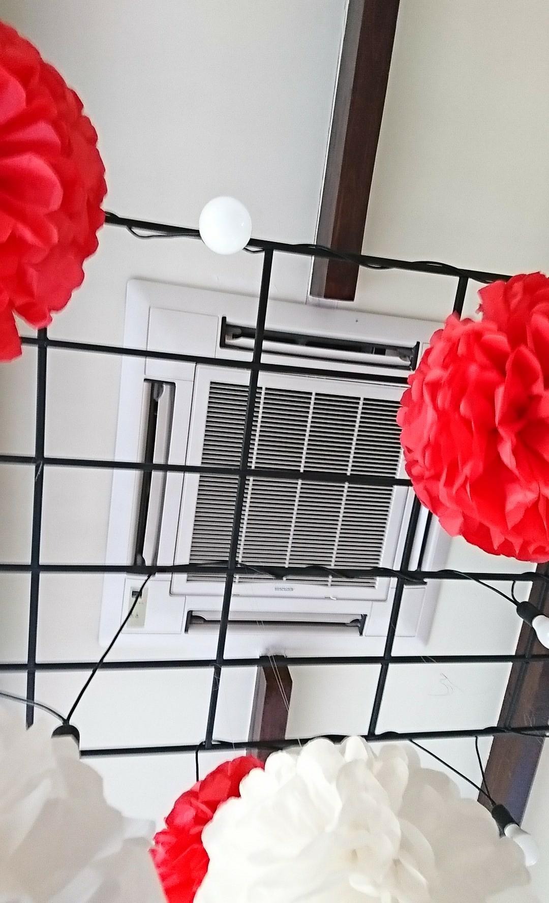 Biała, sufitowa jednostka klimatyzacyjna z widocznymi kratkami wentylacyjnymi, udekorowana czerwonymi i białymi pomponami oraz lampkami, zamontowana pod drewnianą belką.