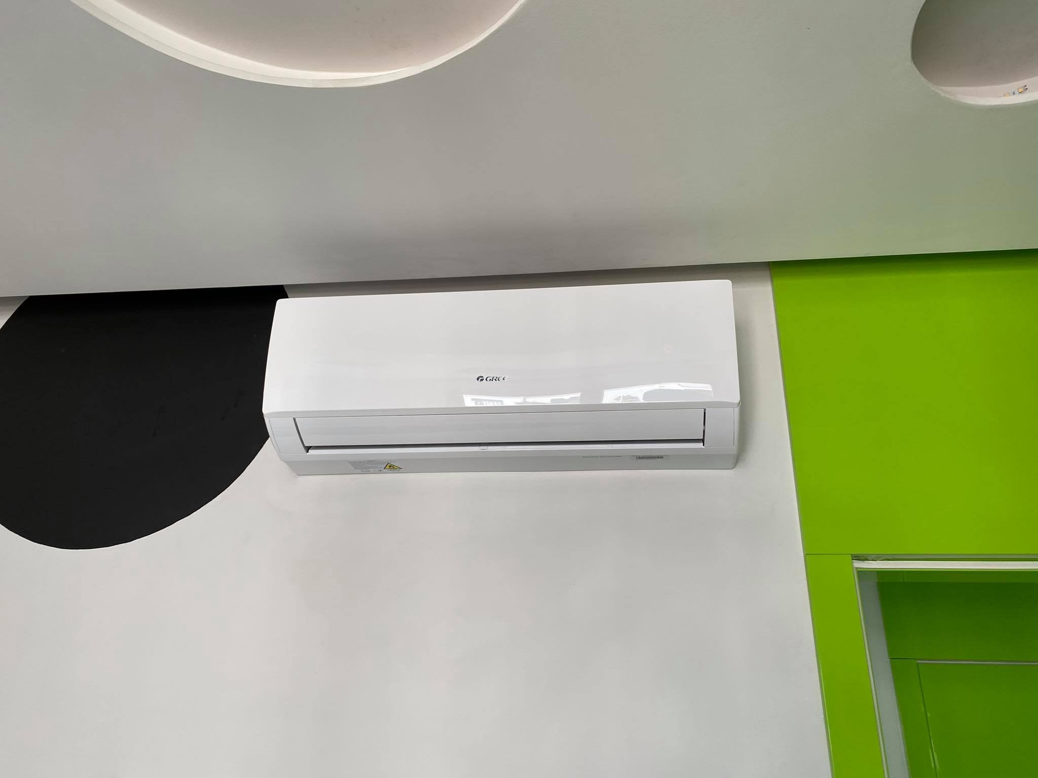 Biała jednostka klimatyzacji marki Gree zamontowana na ścianie z czarno-białym wzorem i jaskrawozielonym elementem architektonicznym.