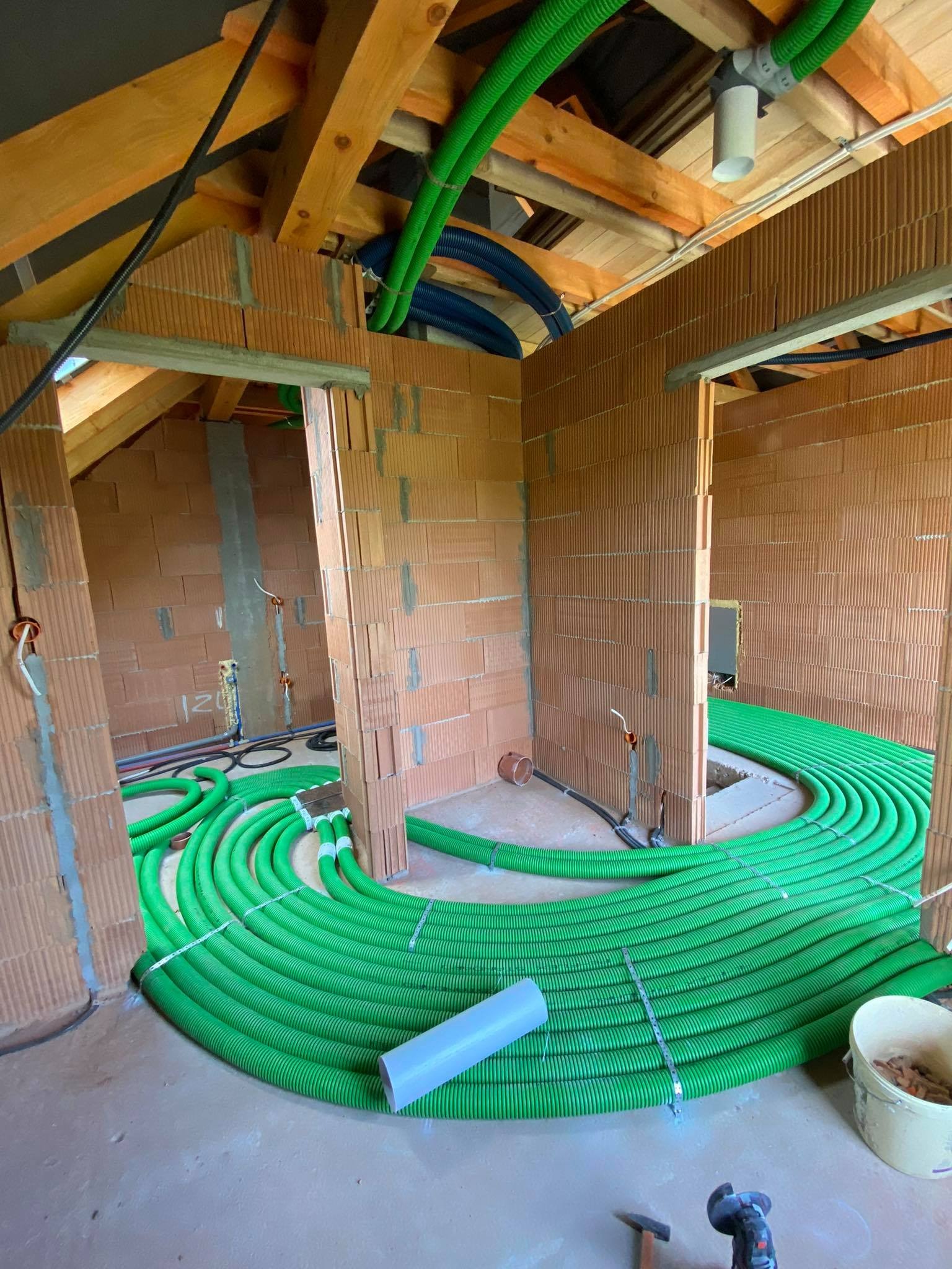 Montaż systemu rekuperacji w surowym stanie budynku: zielone rury rozprowadzone po podłodze, widoczne ceglane ściany i drewniana konstrukcja dachu.