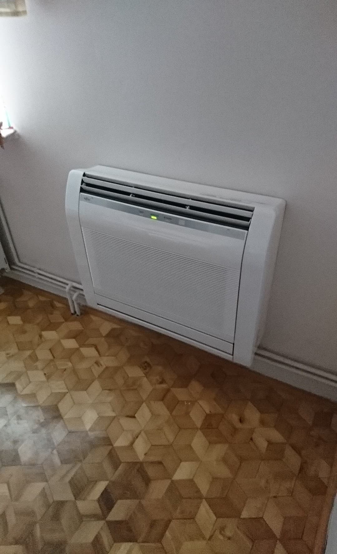 Biały klimatyzator podłogowy Fujitsu zamontowany na ścianie w pomieszczeniu z parkietem w geometryczny wzór. Widoczna zielona dioda sygnalizująca pracę urządzenia.
