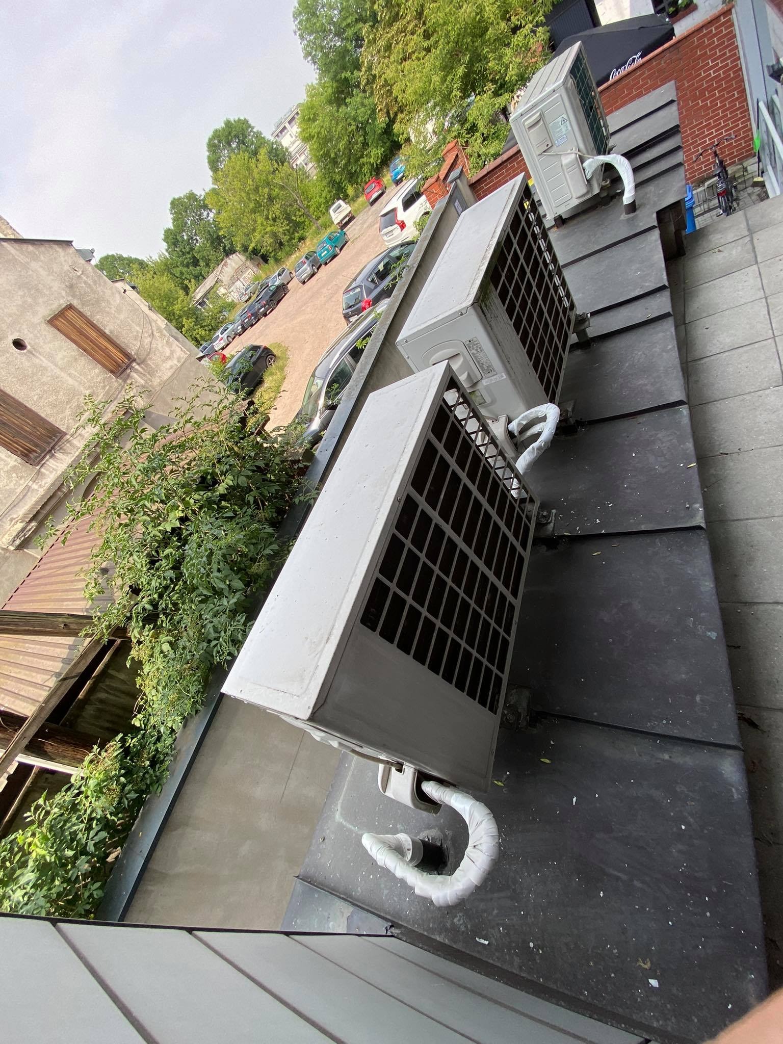 Zewnętrzne jednostki klimatyzacji zamontowane na dachu budynku, widok z góry, z widocznymi przewodami i fragmentem parkingu w tle.