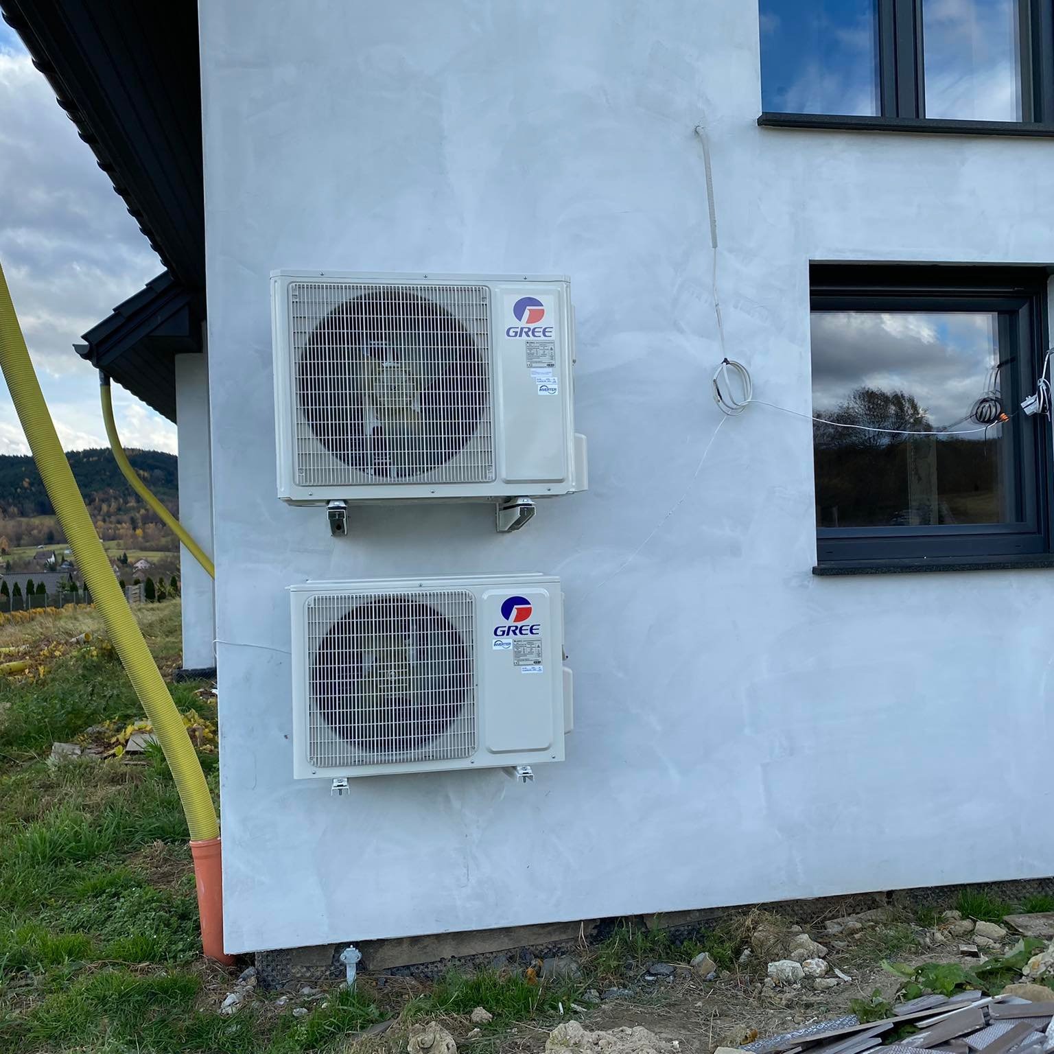 Dwie jednostki zewnętrzne klimatyzacji marki Gree zamontowane na szarej elewacji domu jednorodzinnego z widocznymi przewodami i rurą odprowadzającą skropliny. W tle widoczne wzgórza i fragment nieba.