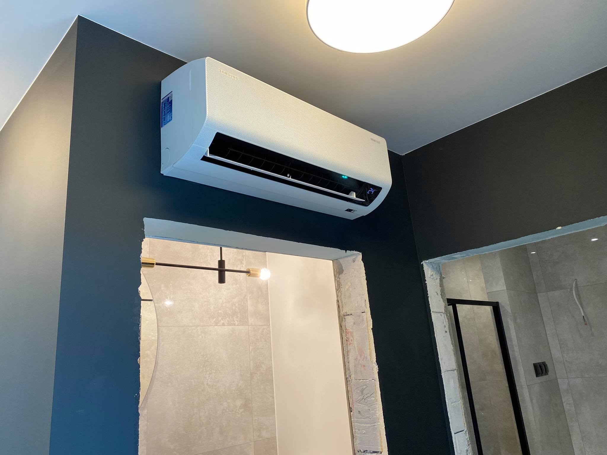 Biała jednostka klimatyzacyjna Samsung zamontowana na ciemnoszarej ścianie, widoczny fragment łazienki z szarą glazurą i czarną ramą kabiny prysznicowej, widoczna temperatura 24 stopnie Celsjusza.