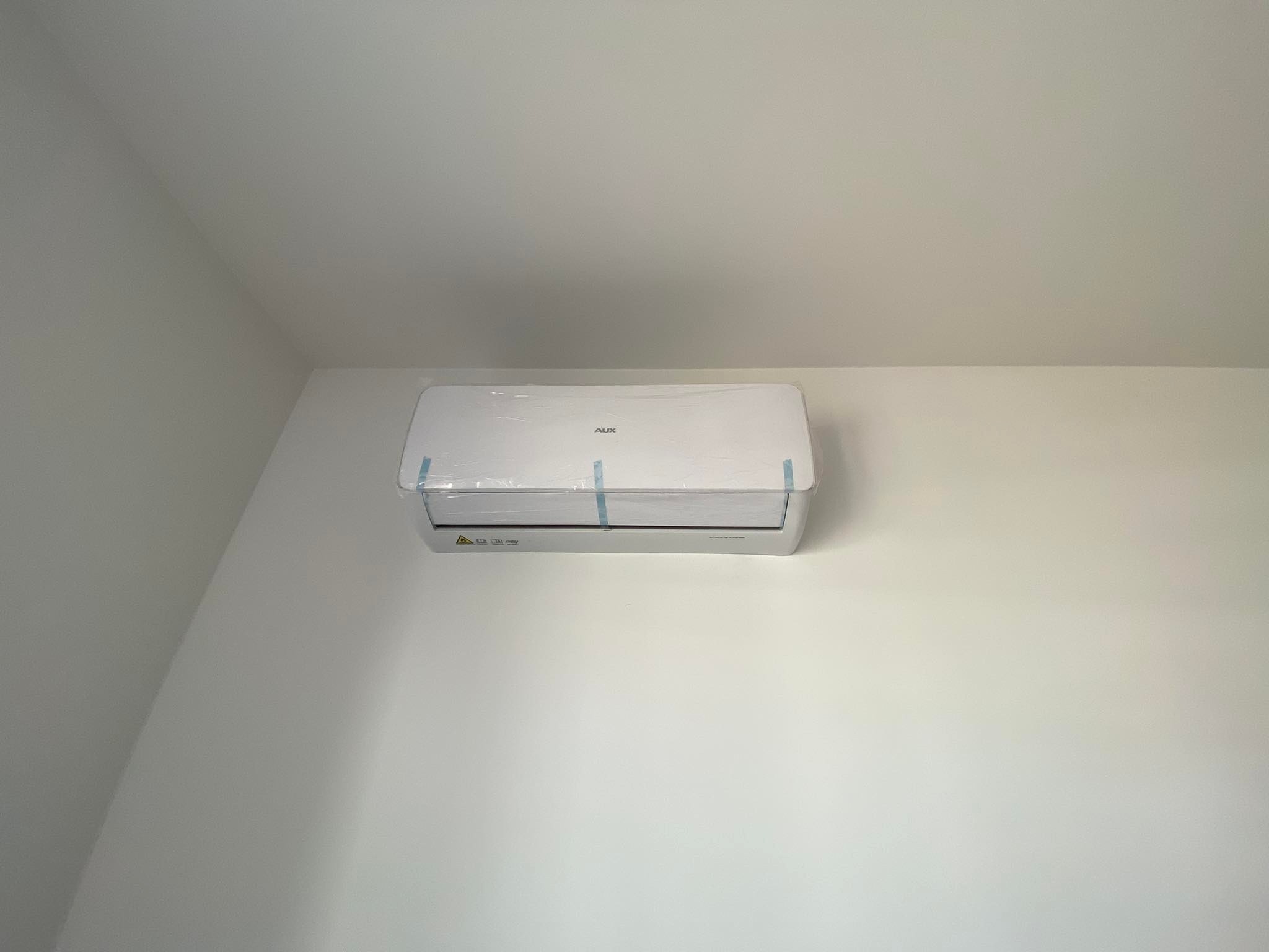 Biała jednostka klimatyzacji AUX, jeszcze w folii ochronnej, zamontowana na białej ścianie w rogu pomieszczenia.