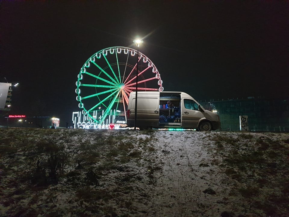 Biały van serwisowy z otwartymi drzwiami, zaparkowany na zaśnieżonym wzgórzu nocą, z podświetlonym diabelskim młynem w tle.