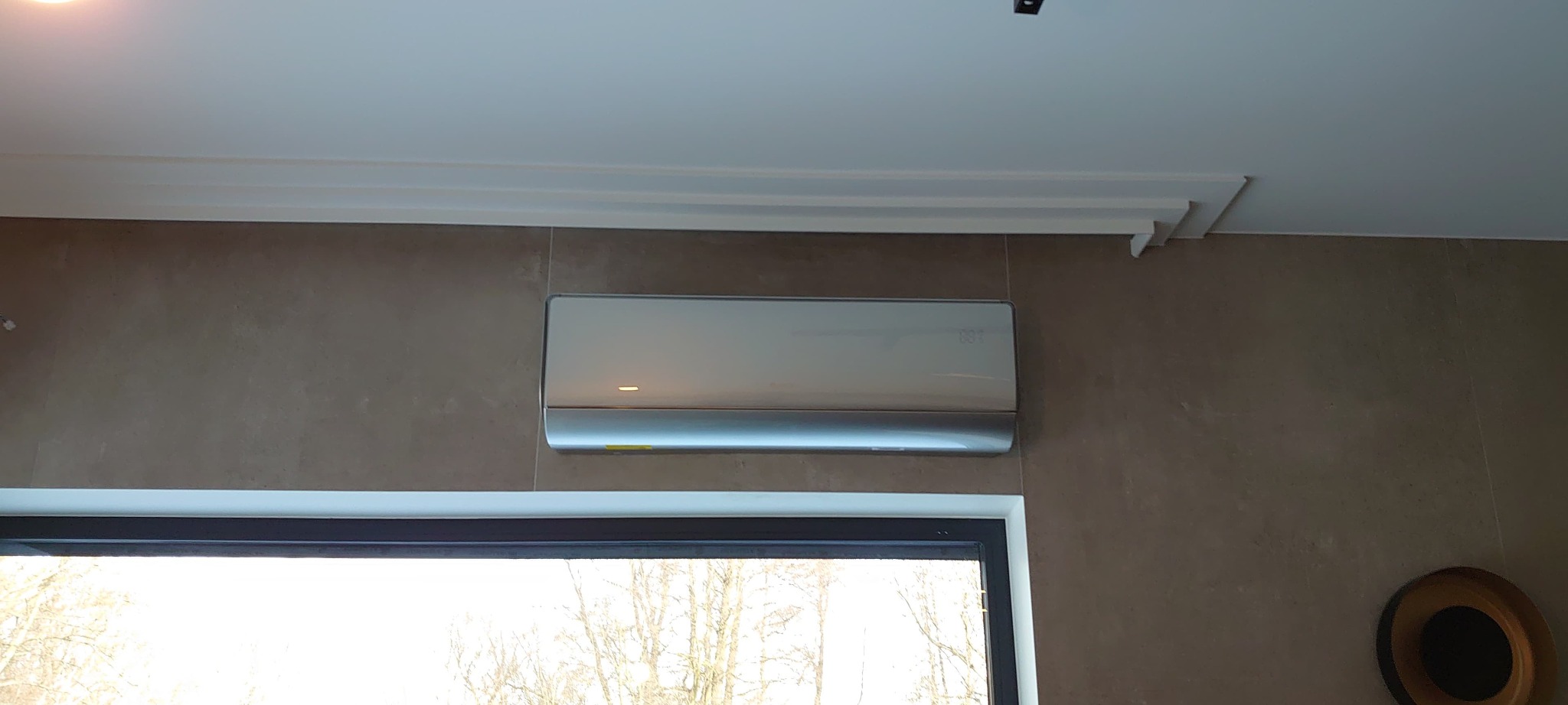 Elegancki, srebrny klimatyzator ścienny zamontowany w nowoczesnym wnętrzu, widok z okna na drzewa bez liści, dekoracyjny gzyms sufitowy.