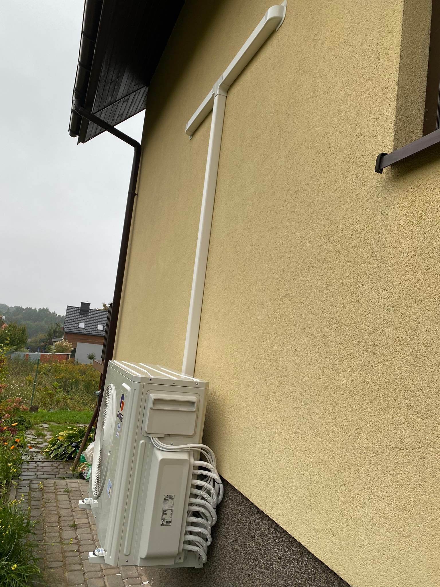 Zewnętrzna jednostka klimatyzacji GREE zamontowana na elewacji domu, z widocznymi przewodami i osłoną odprowadzającą skropliny, na tle pochmurnego nieba i fragmentu ogrodu.