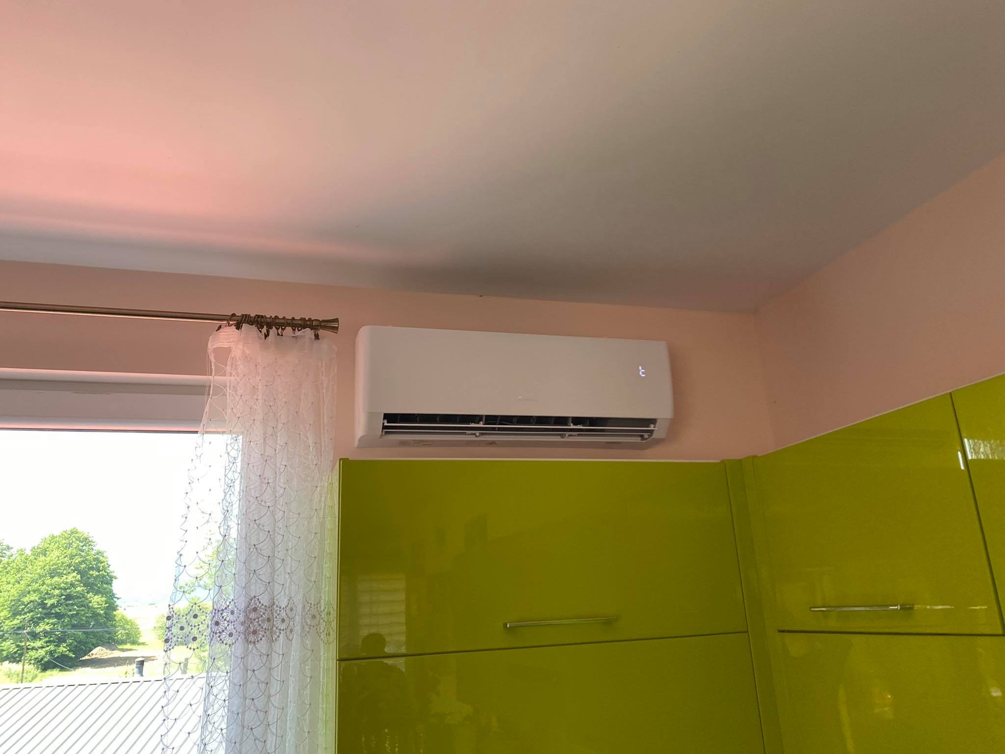 Biały klimatyzator ścienny zamontowany w rogu pokoju z zielonymi szafkami i oknem z firanką