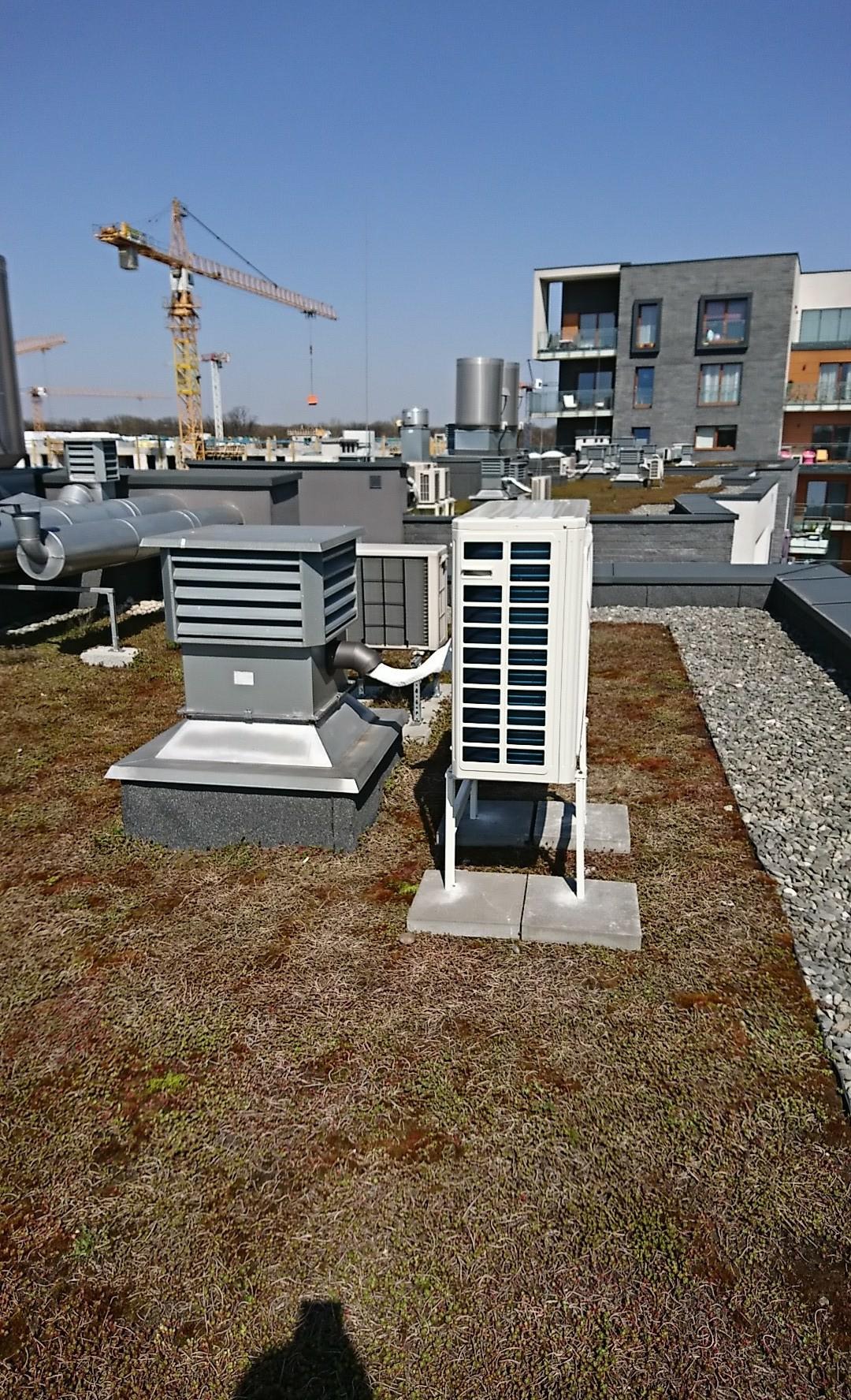Widok z góry na instalacje wentylacyjne i klimatyzacyjne na zielonym dachu budynku komercyjnego z żwirowym obrzeżem, w tle dźwigi budowlane i zabudowa miejska.