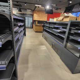 Czyste Miasto - Pusta przestrzeń handlowa z rzędami regałów i chłodziarek przed zapełnieniem towarem, widok z perspektywy podłogi w kierunku zaplecza z ceglaną ścianą.
