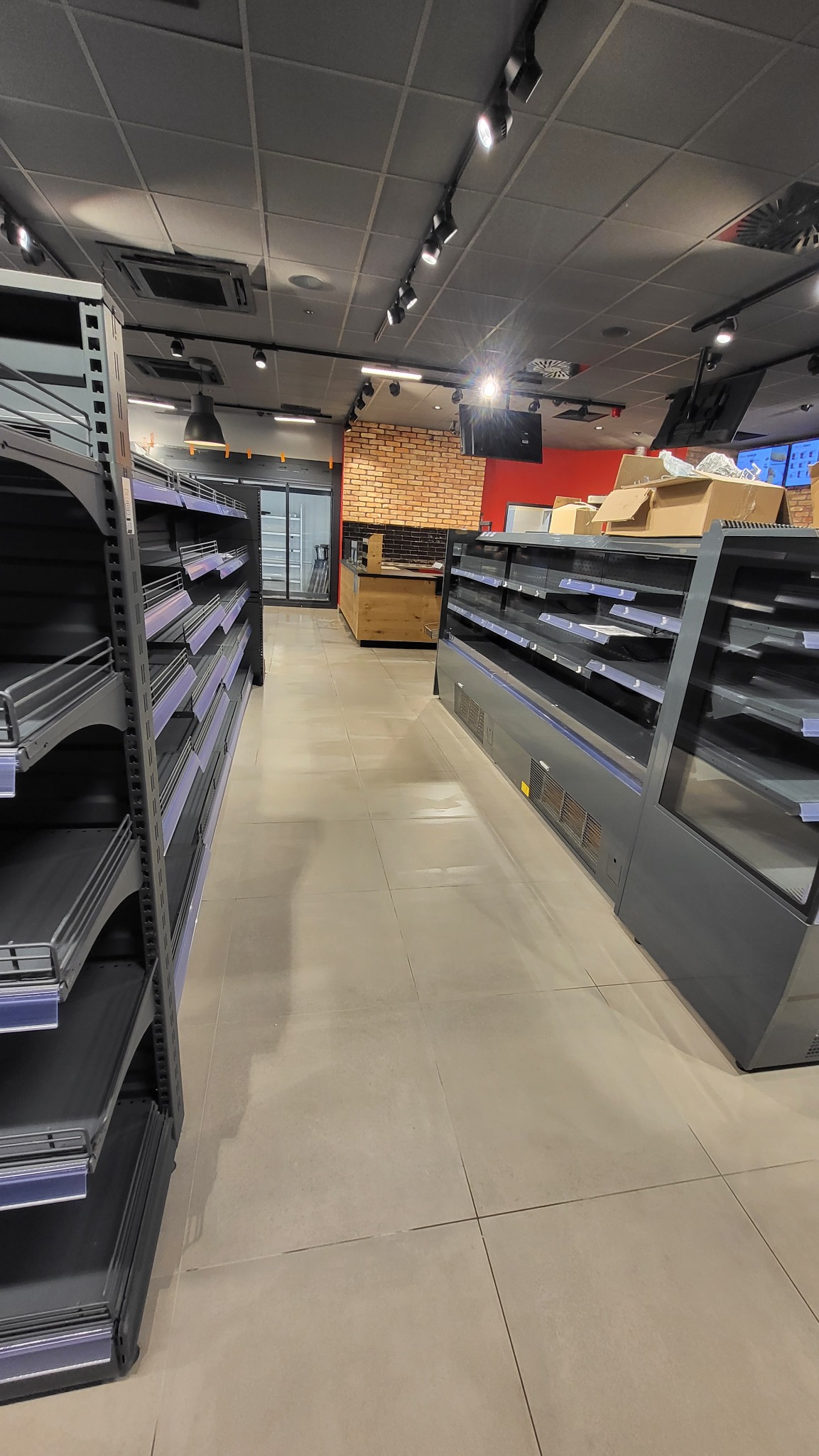 Pusta przestrzeń handlowa z rzędami regałów i chłodziarek przed zapełnieniem towarem, widok z perspektywy podłogi w kierunku zaplecza z ceglaną ścianą.