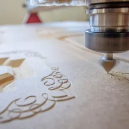 Frezarka CNC wycina ozdobny wzór w płycie drewnianej, widoczne wióry i fragmenty wyciętego elementu.