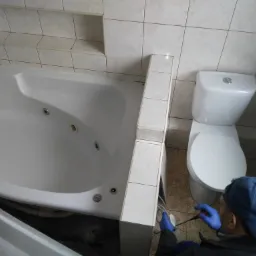 Hydraulik w niebieskich rękawicach i czapce, z narzędziem w dłoni, pracuje w łazience obok wanny z hydromasażem i toalety, prawdopodobnie przy udrażnianiu kanalizacji.